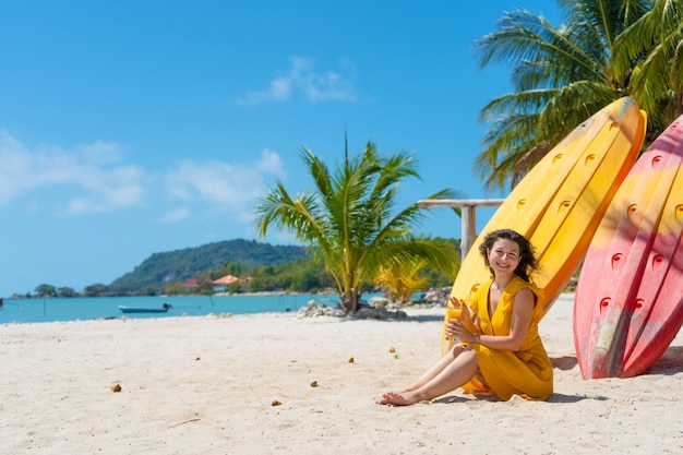 Meisje in een gele jurk op een tropisch zandstrand werkt op een laptop in de buurt van kajaks en drinkt verse mango. Werken op afstand, succesvolle freelance. Werkt op vakantie.