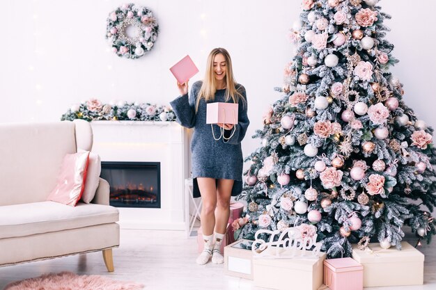 Meisje in een grijze trui opende de geschenkdoos in een gezellige kamer met een open haard, een bank en een kerstboom.