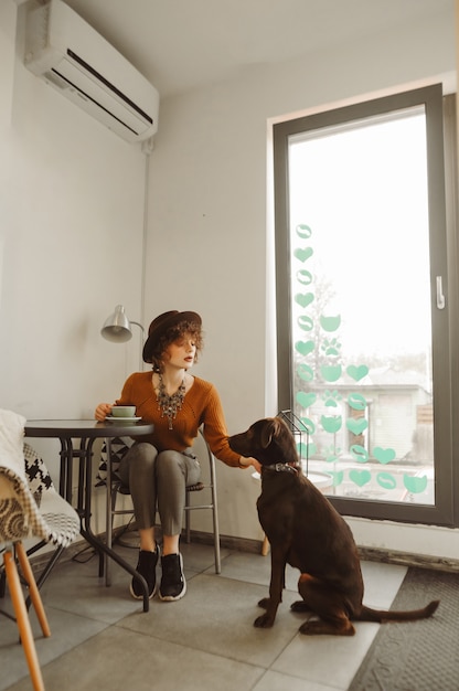 Meisje in een hoed drinkt koffie en speelt met een hond in een gezellig licht café