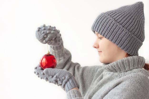 Meisje in een kerstmuts, grijze trui en wanten houdt een rode ballondecoratie voor de kerstboom