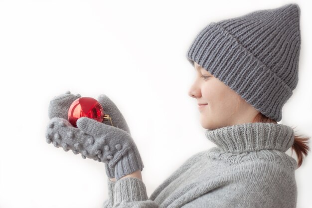 Meisje in een kerstmuts, grijze trui en wanten houdt een rode ballondecoratie voor de kerstboom