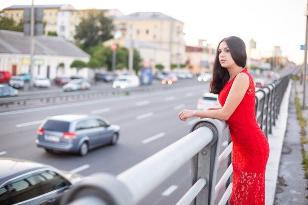 Meisje in een lange rode jurk staat in de buurt van het hek van de rijbaan.