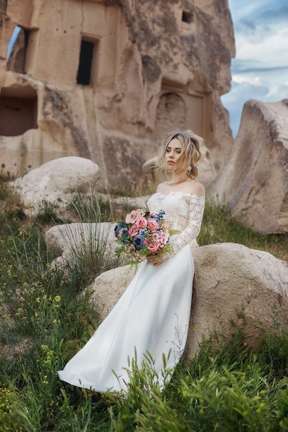 Meisje in een lange trouwjurk met een boeket bloemen in haar handen zit in de bergen in de natuur. Bruiloft in de natuur, relaties en liefde. Bergen van Cappadocië in Turkije