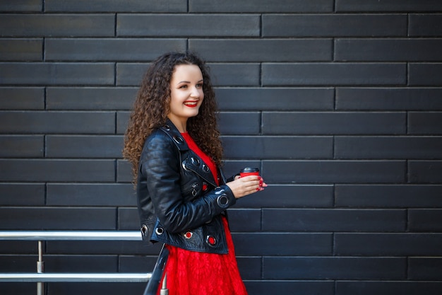 Meisje in een rode jurk en een zwarte jas drinkt koffie uit een rode papieren beker