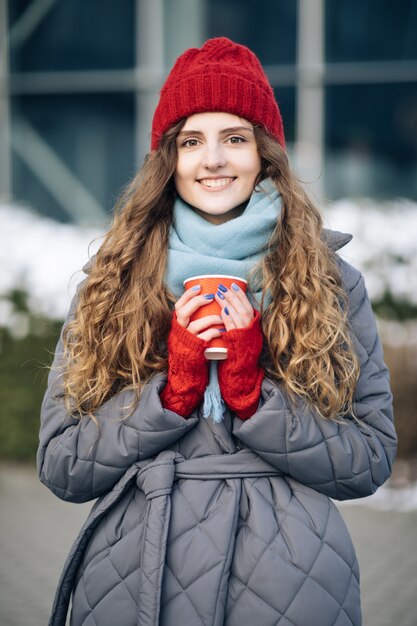 Meisje in een rode warme muts met een kopje koffie in haar handen staat in de winter op straat.