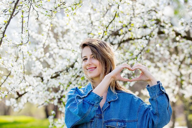 Meisje in een spijkerjasje staat in de buurt van een bloeiende boom en toont harthandteken in het park. lente seizoen