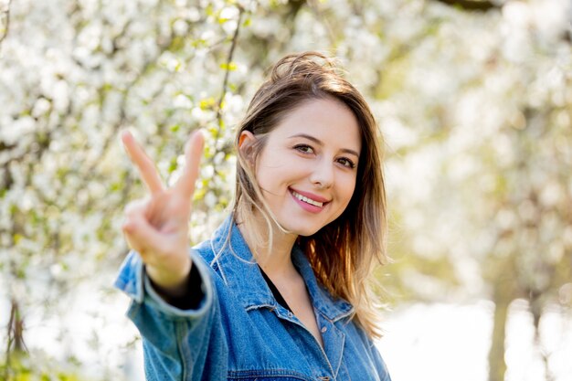 Foto meisje in een spijkerjasje staat in de buurt van een bloeiende boom en toont ok handteken in het park. lente seizoen