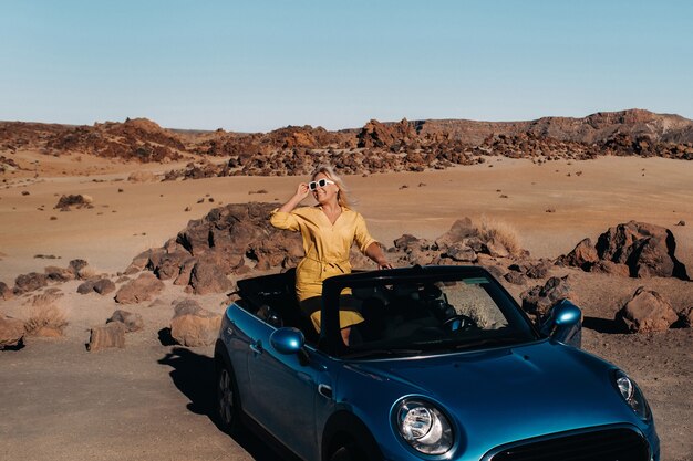 Meisje in gele jurk genieten van een roadtrip in een cabriolet door een verlaten vallei met bergen, Canarische eilanden, Tenerife.