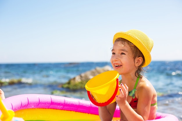 Meisje in gele strohoed speelt met de wind, het water en een waterdispenser in een opblaasbaar zwembad op het strand.