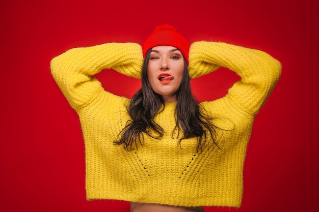 Meisje in gele trui en hoed op rode achtergrond toont tong en knipoogt