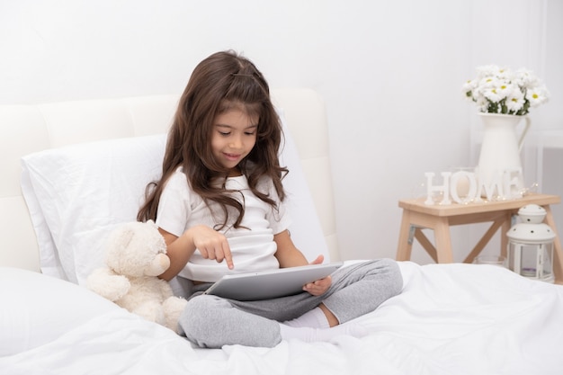 Meisje in huiskleding met teddybeer die tablet zittend op bed gebruikt.