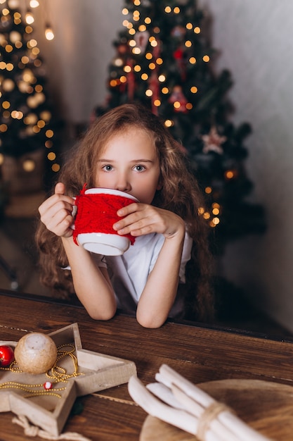 Meisje in Kerstmisdecoratie met thee bij gezellig huis met kleurrijke nieuwe jaarlichten