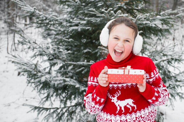 Meisje in Kerstmissweater en oorkappen met gift in winterbos