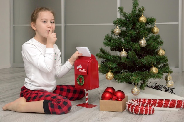 Meisje in pyjama bij de kerstboom gooit een brief aan de kerstman in de kerstcadeaus van de brievenbus