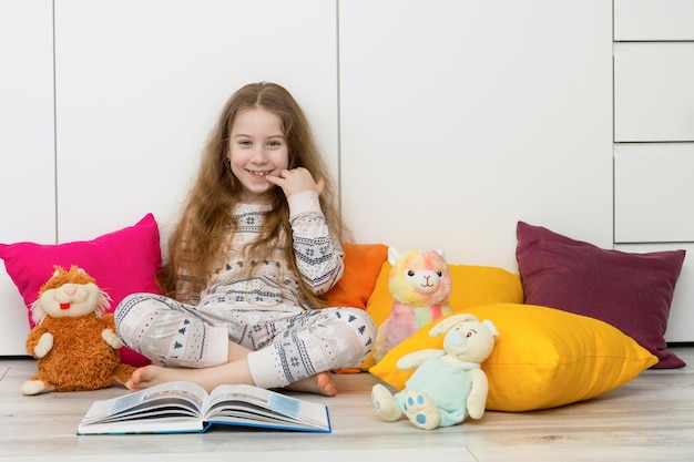 Meisje in pyjama zit op de vloer tussen kleurrijke kussens en een open boek en lacht vrolijk