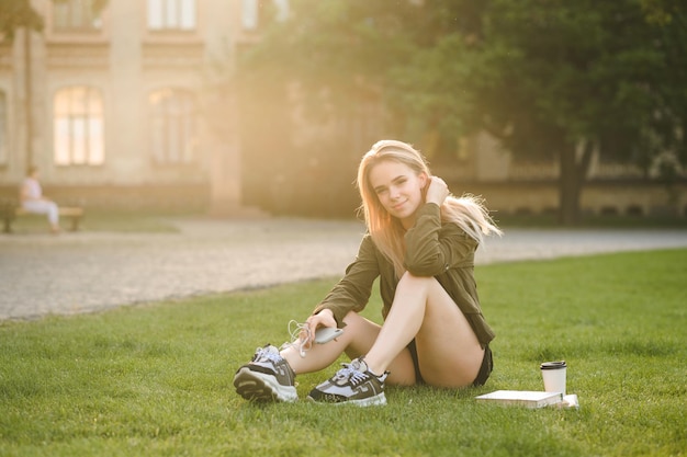 meisje in stijlvolle vrijetijdskleding zit op het grasveld tegen de achtergrond van een universiteitsgebouw