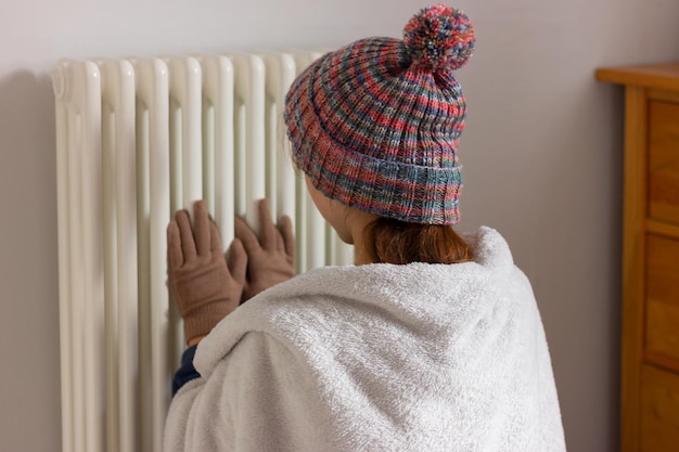 Meisje in warme kleren en een hoed in de buurt van de verwarmingsradiator in de kamer, verwarming, warmtebehoud en besparingen