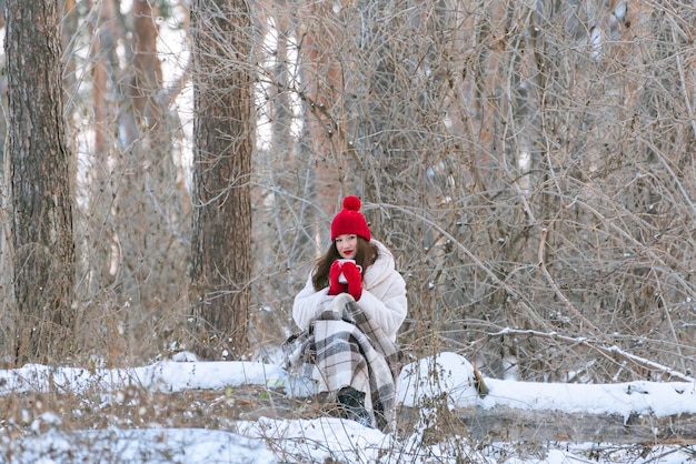 Meisje in winter forest drinkt thee gewikkeld in deken. Vrouw in rode hoed in besneeuwde pack. Ruimte kopiëren