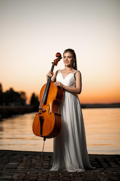 Meisje in witte mooie jurk met een cello staat bij het meer bij zonsondergang