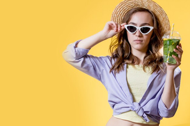 Meisje in zomeroutfit met witte trendy zonnebril en genietend van een mojito-cocktail. poseren op de gele achtergrond.