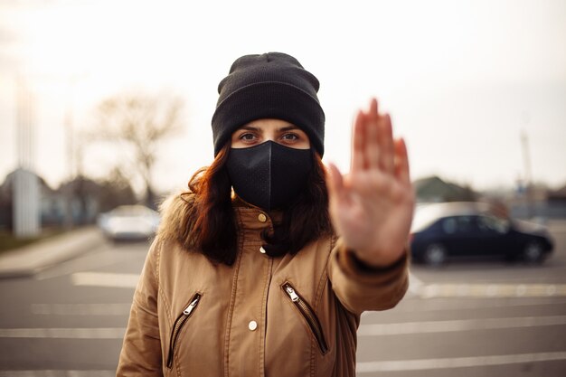 Meisje, jonge vrouw in beschermend steriel medisch zwart masker op haar gezicht camera buitenshuis kijken, op straat toont palm, hand, stop geen teken. Vervuiling, virus, pandemisch coronavirus concept.