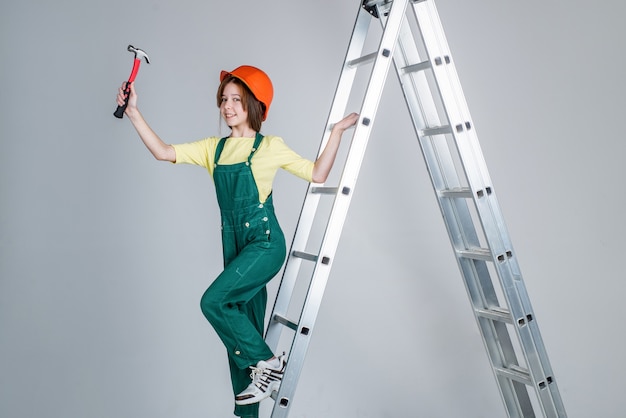 Meisje kind dragen uniform en helm tijdens het werken in werkplaats met hamer op ladder, carrière.