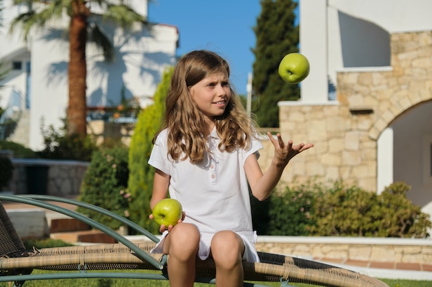 Meisje kind gooien, spelen, jongleren met groene appels, zittend op een stoel buiten op groen gazon