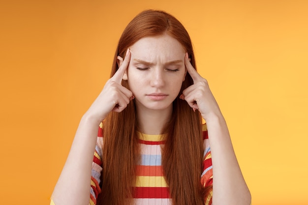 Meisje krijgt gedachten opgestapeld en probeert helder te denken concentreren zich belangrijke taak te concentreren onthoud nummer aanraken tempels sluiten ogen kijken serieus staand verbaasd lijden aan hoofdpijn, oranje achtergrond