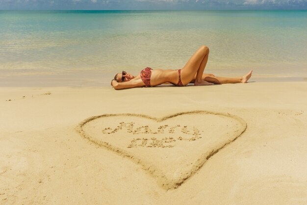 Meisje liggend op het zand in het midden van het strand met een hart getekend op het zand met de tekst merry me in het midden