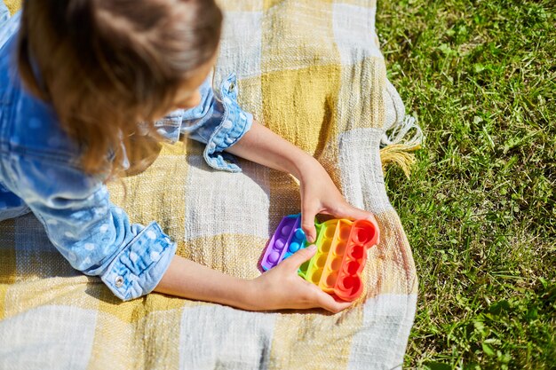 Meisje ligt buiten op een deken op gras en speelt pop it, kid hands spelen met kleurrijke pop it, fidget speelgoed in de achtertuin van het huis op een zonnige zomerdag, zomervakantie.