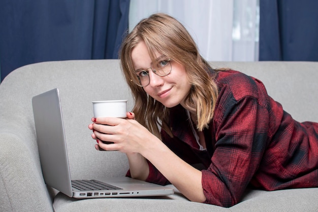 Meisje ligt op de bank met een laptop en kijkt naar een video die een vrouwelijke freelancer thuis werkt