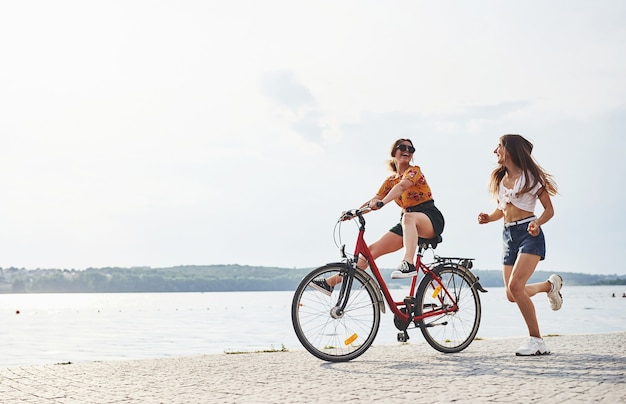 Meisje loopt in de buurt van fiets. Twee vriendinnen op de fiets hebben plezier op het strand bij het meer.