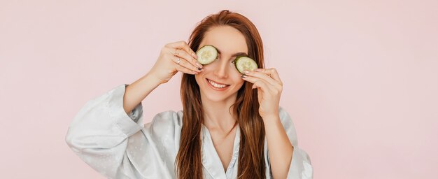 Meisje maakt zelfgemaakte gezichtsmaskers. Komkommers voor de frisheid van de huid rond de ogen. Vrouw zorgt voor een jeugdige huid. Model lachen en plezier hebben in de spa op roze achtergrond