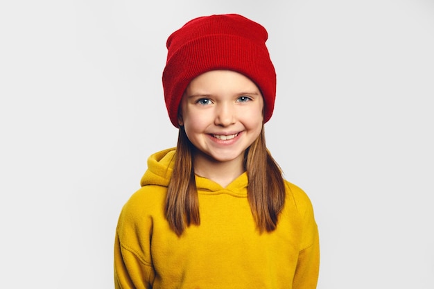 Meisje met aangename glimlach draagt gele hoodie en rode hoed staat tegen