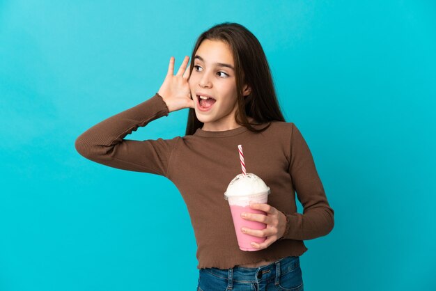 Meisje met aardbei milkshake geïsoleerd op blauwe achtergrond luisteren naar iets door hand op het oor te leggen