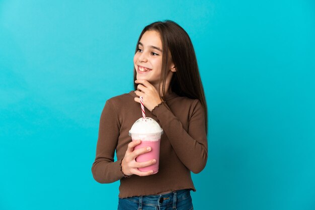 Meisje met aardbei milkshake geïsoleerd op blauwe achtergrond op zoek naar de kant en glimlachen