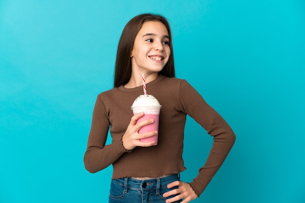 Meisje met aardbei milkshake geïsoleerd op blauwe achtergrond poseren met armen op heup en glimlachen