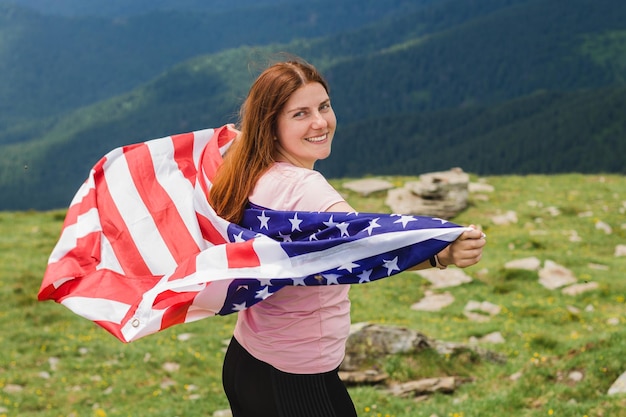 Foto meisje met amerikaanse vlag die uitkijkt op landschap jonge vrouw staat in de bergen met de amerikaanse vlag in haar armen hoog vlag wappert in de wind