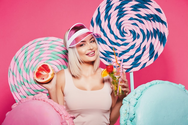 Meisje met blond haar met een top en een roze pet die zich met enorme zoete lolly's op roze bevindt