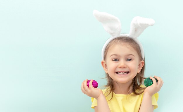 meisje met bunny oren hoofdband en gele jurk op blauwe achtergrond houdt gekleurde eieren