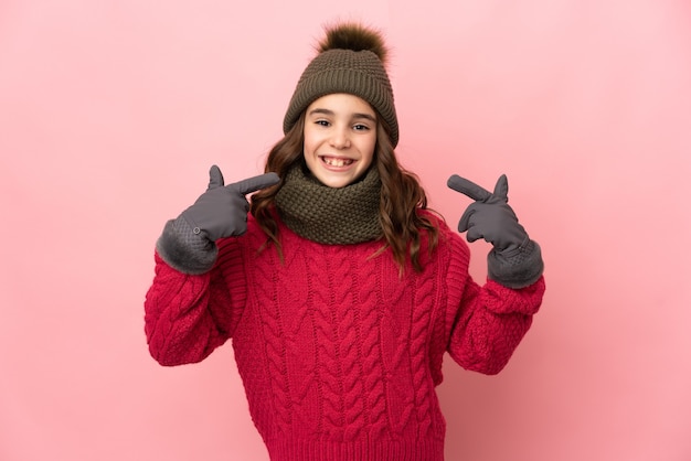 Meisje met de winterhoed die op roze achtergrond wordt geïsoleerd die een duim omhoog gebaar geeft