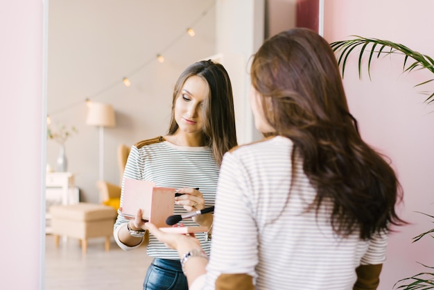 Meisje met donker haar staat voor een spiegel en doet make-up met een penseel in haar hand en creëert een schoonheid en stijl van het beeld