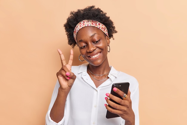meisje met donkere huid krullend Afro haar maakt vredesgebaar neemt selfie of maakt video-oproep via smartphone draagt hoofdband wit overhemd geïsoleerd op beige hebben. Technologie concept