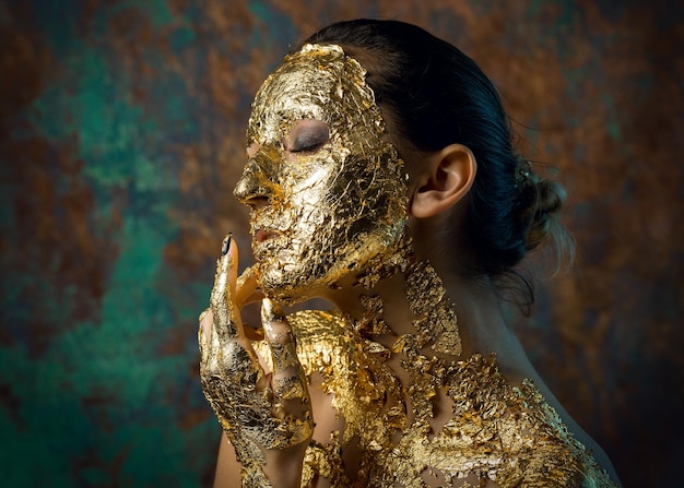 Meisje met een masker op haar gezicht gemaakt van bladgoud Somber studioportret van een brunette op een abstracte achtergrond