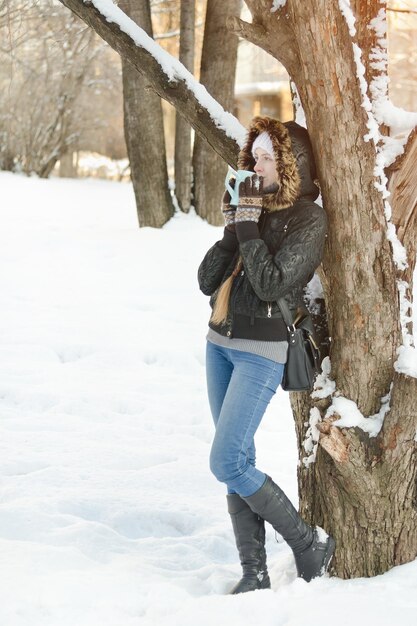 Meisje met een mok thee leunend tegen een boom en recht kijkend Winter buitenshuis