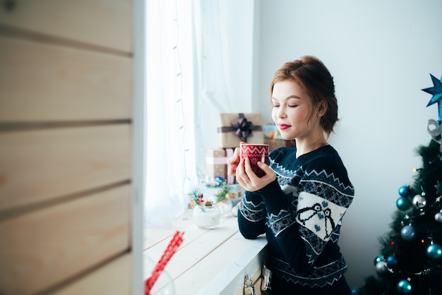 Meisje met een rode mok in haar handen bij het raam, op de achtergrond van een kerstboom