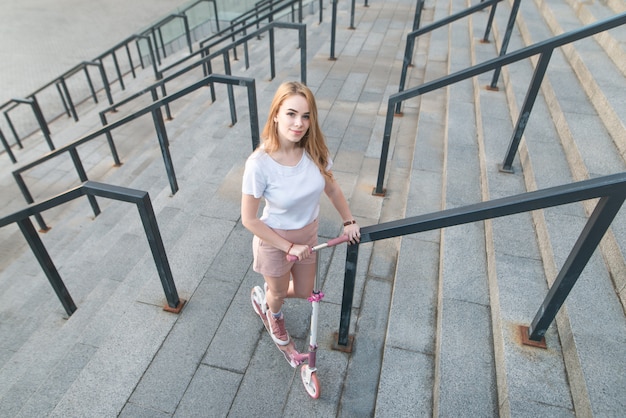 Meisje met een roze kick-scooter staat op de achtergrond van een moderne architectuur, weergave van de camera, bovenaanzicht.