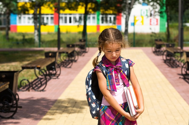 Meisje met een rugzak en een stapel boeken in de buurt van de school. Terug naar school, het kind is moe, zware leerboeken. Onderwijs, basisschoolklassen, het begin van het schooljaar, 1 september