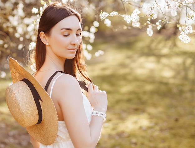 Meisje met een strohoed in de lente in het park Brunette met lang haar houdt een hoed vast op een achtergrond van zomerse natuur Jeugd en schoonheid