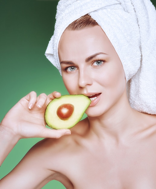 Meisje met een witte handdoek op haar hoofd met een voedzaam groen masker op haar gezicht en een avocado in haar handen op een groene achtergrond met ruimte voor tekst